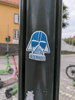 Street sticker The sticker features a stylized, simplified depiction of Darth Vader's helmet in blue and white. Below the helmet, the word "HEISENVADER" is written in a blocky, somewhat pixelated font. The design merges elements of the iconic Star Wars character with a nod to the Breaking Bad series, specifically Walter White's alter ego, Heisenberg. The sticker is placed on a dark, possibly metallic, pole outdoors, with blurred elements of a street scene, buildings, and scooters in the background.