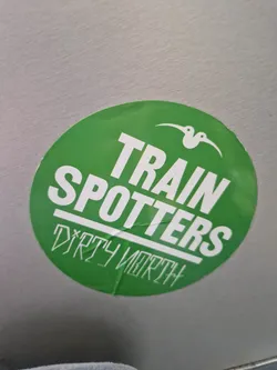 Street sticker Round green sticker with the text "TRAIN SPOTTERS" in white bold uppercase letters. Underneath, in a smaller, stylized font, it says "DIRTY NORTH". A small bird-like logo is present at the top.