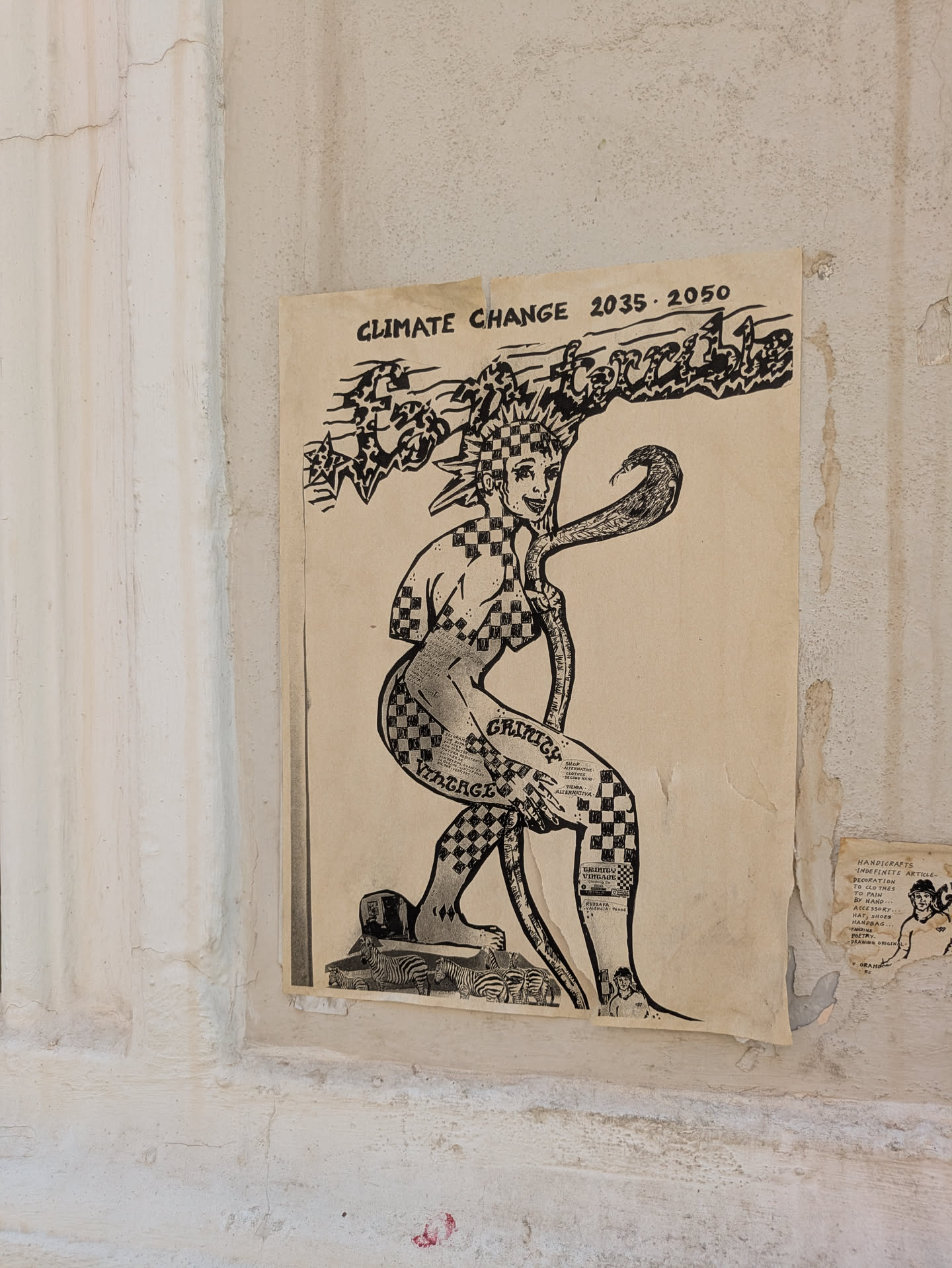 The image shows a black and white poster attached to a wall. The poster presents an illustration of a female figure with a snake draped around her neck. The figure and snake have a checkerboard pattern. Text on the poster reads "CLIMATE CHANGE 2035-2050" and "GRINICY VINTAGE". The overall theme seems to address climate change in a creative and artistic way.