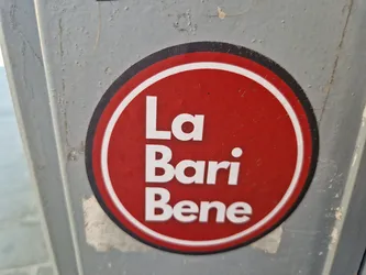 A red and white circular sticker with the text "La Bari Bene" in a simple sans-serif font. The sticker is affixed to a gray surface.