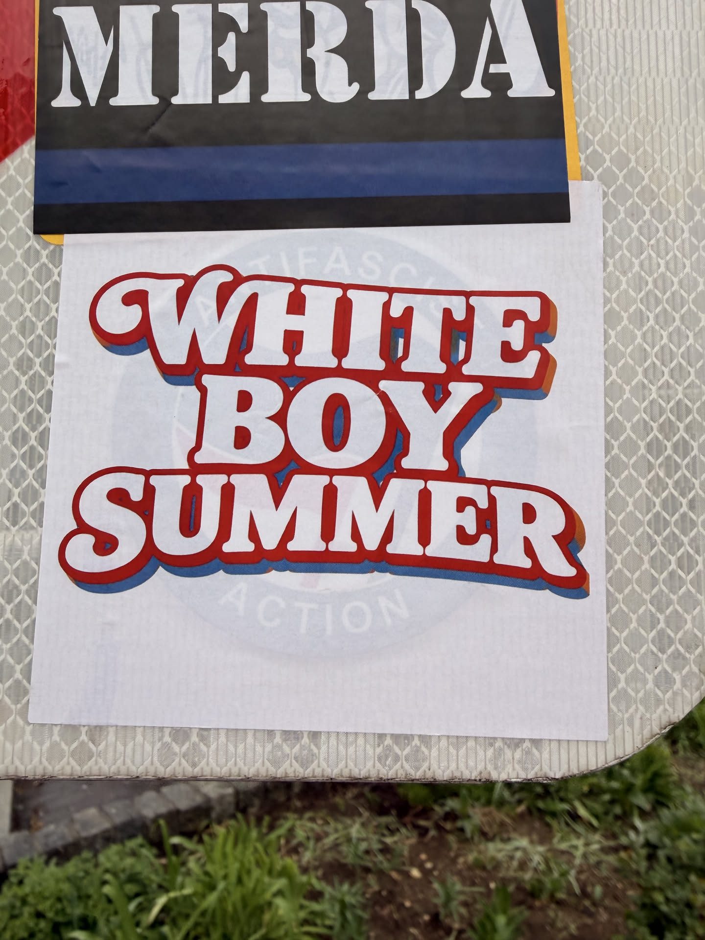 The sticker is a white rectangular decal featuring the phrase "WHITE BOY SUMMER" stacked in three lines. The text is rendered in a prominent, rounded, sans-serif font with a distinct retro or vintage style. Each letter is white, outlined in a thick red, followed by a thinner orange drop shadow, and a final blue drop shadow, creating a vibrant, multi-layered, and three-dimensional effect. Faintly visible in the background, beneath the main text, is a light blue circular graphic with some barely legible text, which appears to be 'ANTIFA' and 'ACTION', but it is largely obscured and faded.