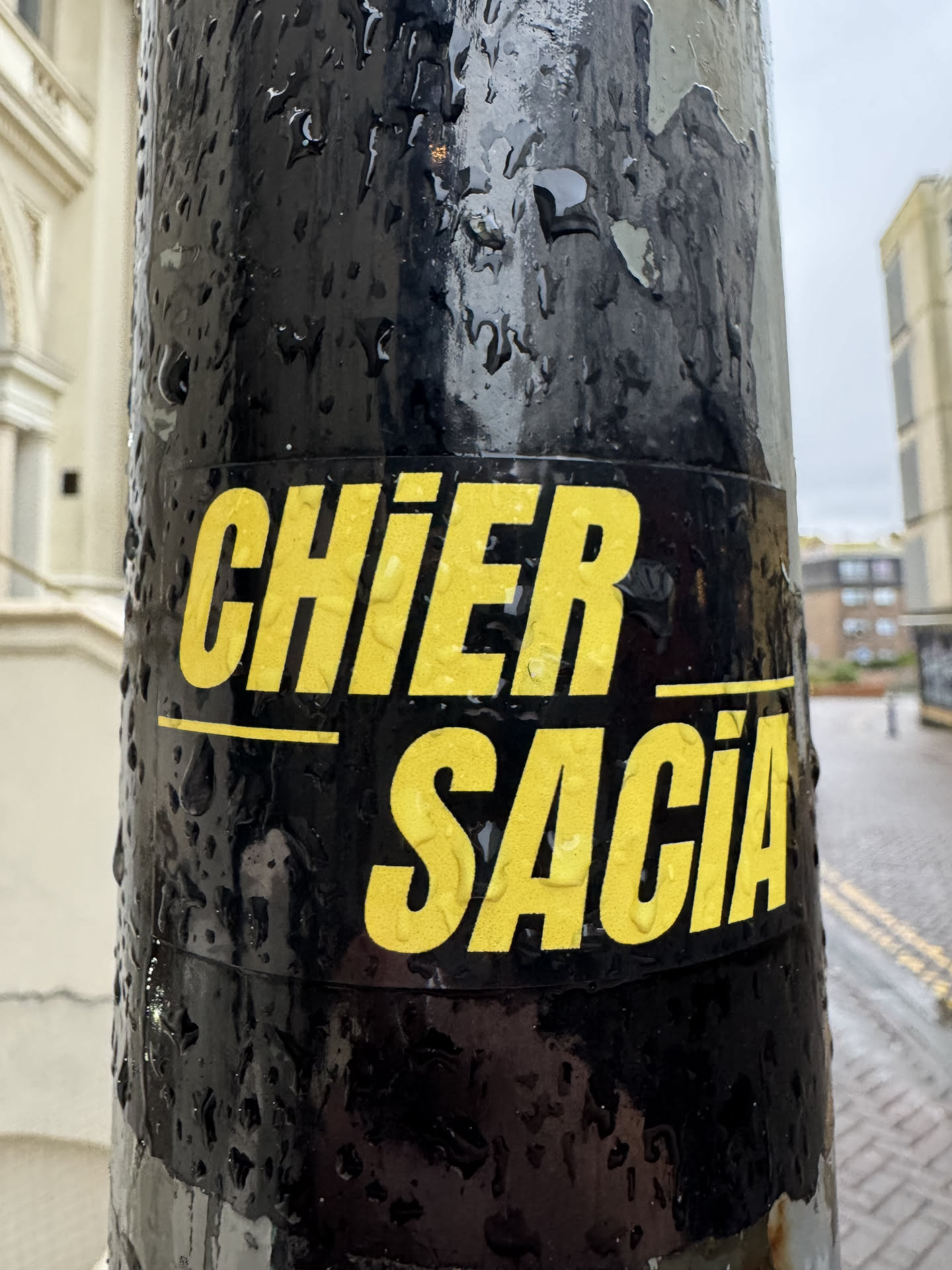 Street sticker A rectangular sticker featuring two lines of bold, sans-serif yellow text on a solid black background. The top line reads 'CHIER' and the bottom line reads 'SACIA'. A thin yellow horizontal line separates the two words. The sticker is applied to a wet, black pole, with visible raindrops clinging to its surface and the pole itself. The text has a slightly textured appearance due to the rain.