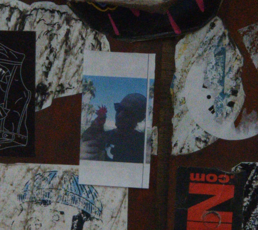 The sticker is a rectangular image with a white border, resembling an instant photo print. The central image features a dark, almost entirely black silhouette of a person, likely wearing a baseball cap or similar headwear. The person is depicted from the chest up, facing slightly to the right. Their right hand is raised, holding a small, bright red object, which could be a flower, fruit, or another small item. The background is a clear, bright blue sky, with faint, blurry hints of green foliage visible on the left side, suggesting trees or bushes. The overall aesthetic is simple and focuses on the contrast between the dark silhouette and the bright background, with the red object providing a single point of color.