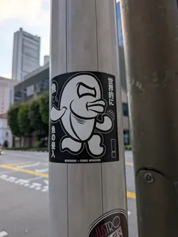 A rectangular black sticker depicting a stylized, walking cartoon fish character with an expressive face. The design uses bold white outlines on a black background, accompanied by vertical Japanese text on the sides and English text at the bottom.