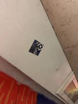 Street sticker The sticker is a diamond-shaped graphic with a dark blue border. It features an illustration of a male figure, likely a football player, with dark, curly hair, wearing a dark blue and white striped or patterned jersey. His right arm is raised in a celebratory gesture, with a clenched fist. The word "HIDACH" is printed in white on the front of the jersey. To the right of the figure, a prominent white diamond logo with a black outline is visible against a blue background, strongly resembling the Hamburger SV (HSV) football club emblem. The overall aesthetic is reminiscent of vintage sports memorabilia, with a slightly worn appearance.