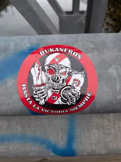 The sticker is circular with a prominent red border. At its center is a stylized skull with glowing red eyes, wearing a white and red striped bandana or helmet with a 'K' emblem. The skull is depicted with sharp teeth and a menacing grin. In its right hand, it holds a dagger with a lightning bolt design on the blade. In its left hand, it holds a lit bomb with a fuse and an explosion graphic. The skull is also wearing a white shirt with a red tie and a small emblem. The text 'BUKANEROS' is written in white capital letters along the top inner edge of the red border, and 'HASTA LA VICTORIA SIEMPRE' is written in white capital letters along the bottom inner edge of the red border.