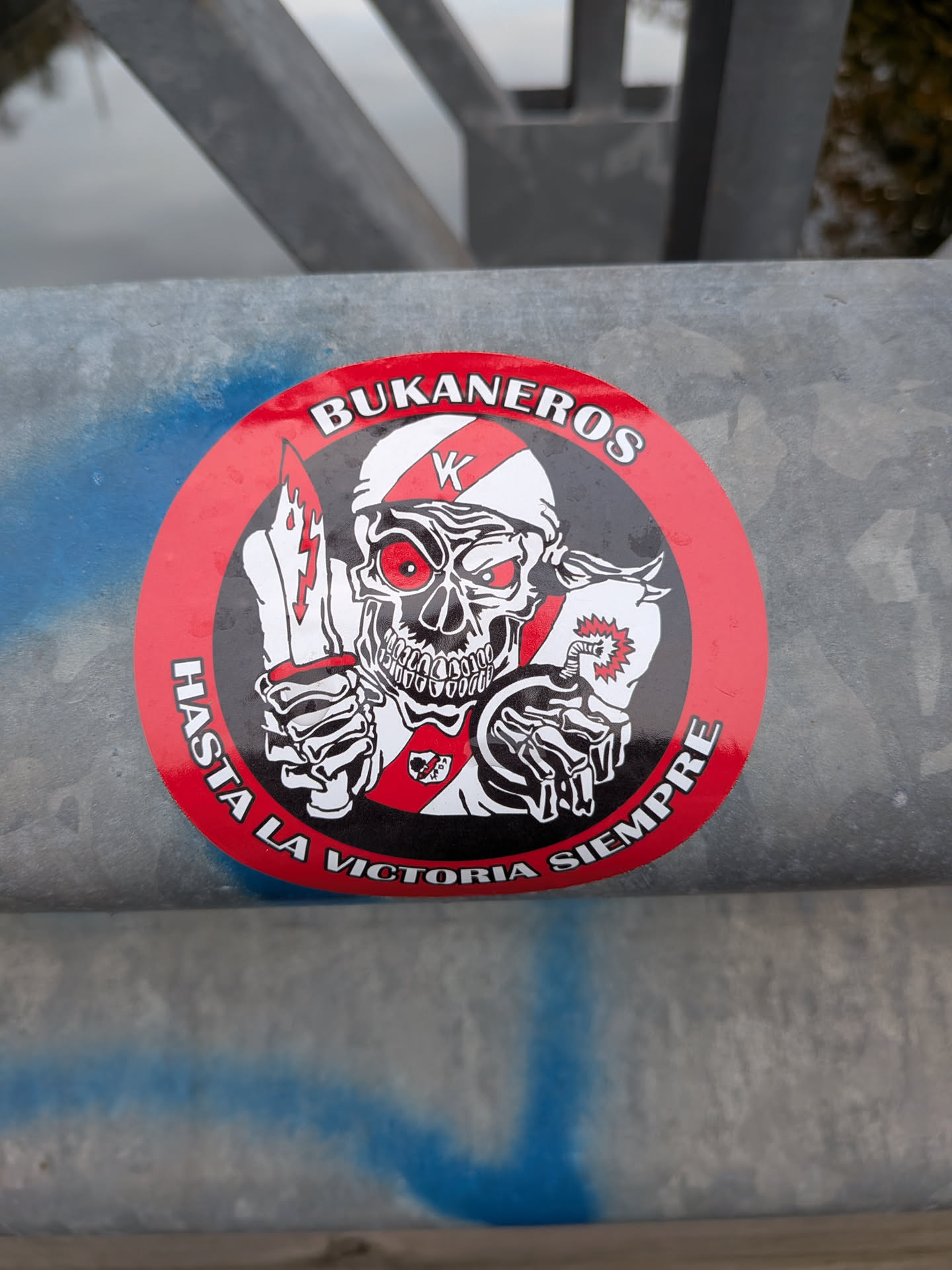 Street sticker The sticker is circular with a prominent red border. At its center is a stylized skull with glowing red eyes, wearing a white and red striped bandana or helmet with a 'K' emblem. The skull is depicted with sharp teeth and a menacing grin. In its right hand, it holds a dagger with a lightning bolt design on the blade. In its left hand, it holds a lit bomb with a fuse and an explosion graphic. The skull is also wearing a white shirt with a red tie and a small emblem. The text 'BUKANEROS' is written in white capital letters along the top inner edge of the red border, and 'HASTA LA VICTORIA SIEMPRE' is written in white capital letters along the bottom inner edge of the red border.