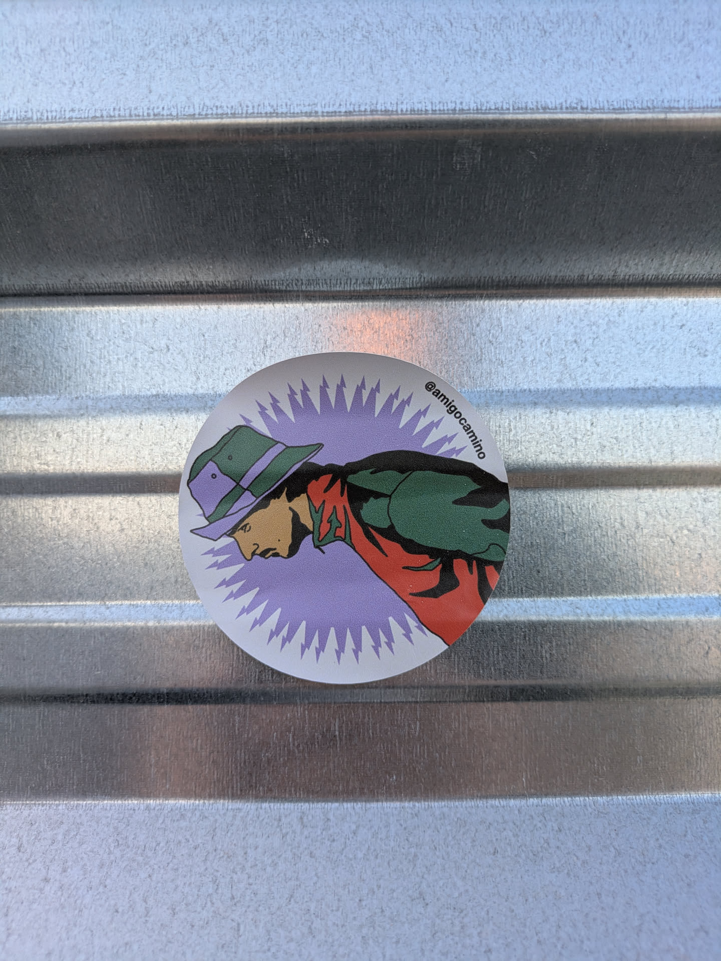 This round sticker features a character illustration with a green and purple hat, a green jacket, and a red scarf, all drawn in a comic book style. The background is a vibrant purple with radial lines that suggest energy or movement. The text '@amigocamino' is present, indicating a possible social media handle.