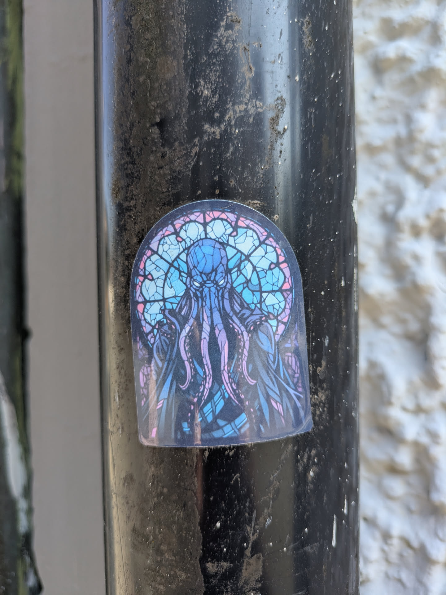 The sticker features an intricate and vibrant illustration of a mythical deep-sea creature, resembling an octopus or kraken, rendered in a stained-glass mosaic style. The creature has a prominent head, multiple winding tentacles, and piercing eyes. The design uses a cool color palette dominated by various shades of blue, purple, and hints of pink, all outlined with dark lines, creating the segmented look of leaded glass. The sticker itself is shaped with an arched top, mimicking a decorative window. It is adhered to a dirty, dark grey or black metal pole.