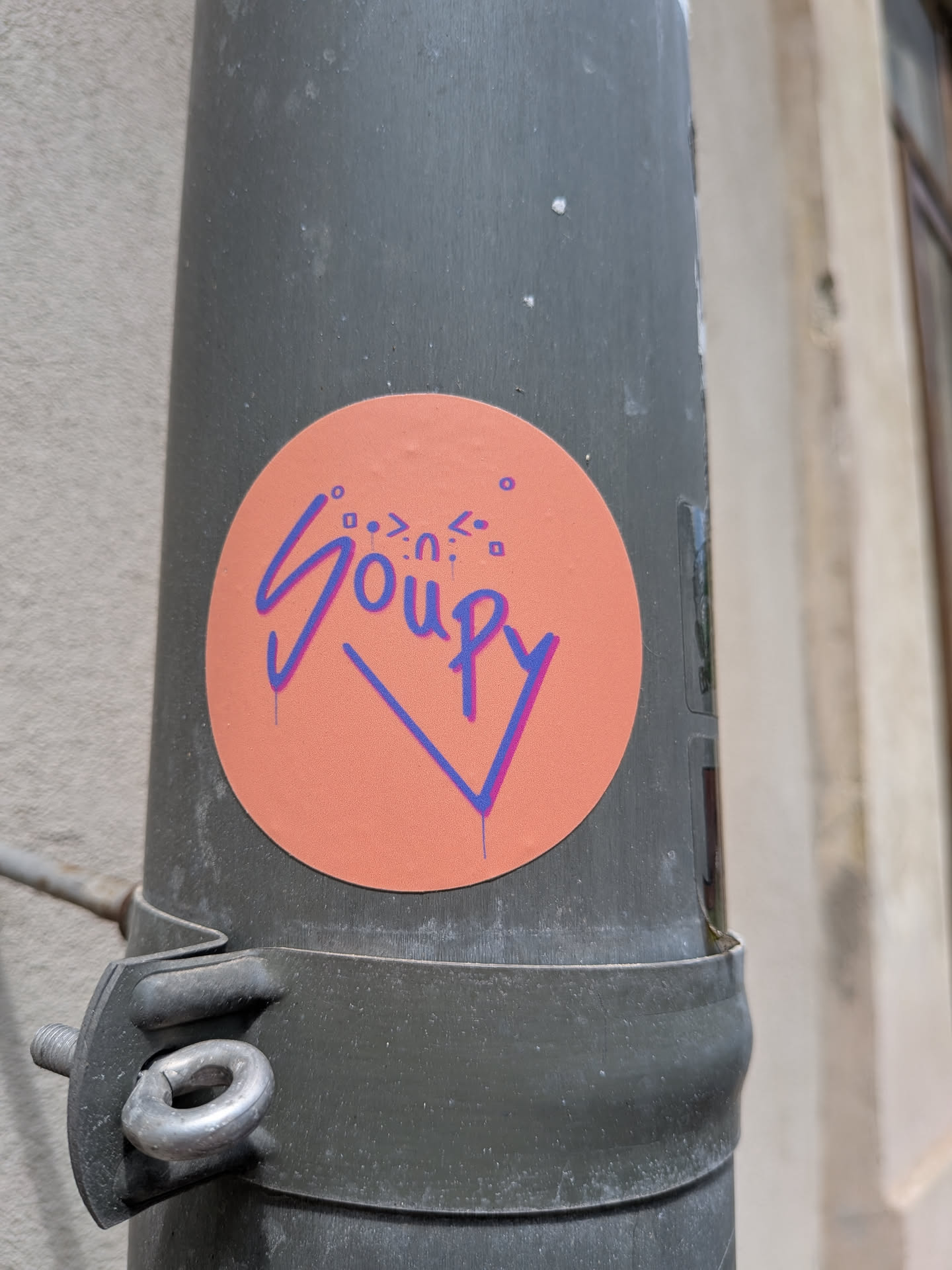 A round peach-colored sticker on a grey pole. It displays the word 'Soupy' in a blue and purple graffiti-style font, topped with small abstract symbols.