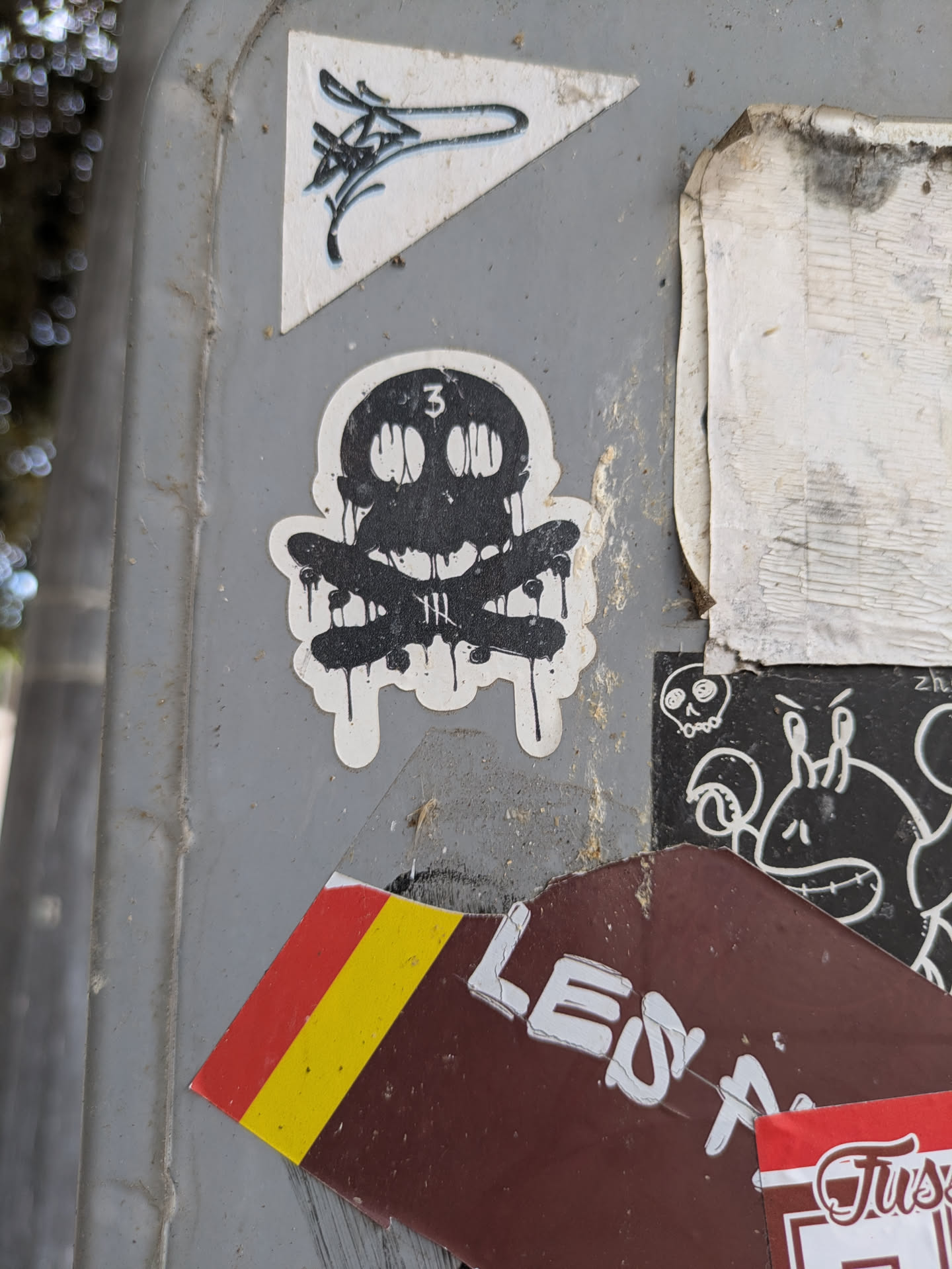 A black and white sticker showing a dripping skull and crossbones is adhered to a light pole among other stickers. The design is simple and bold.