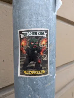 Street sticker The sticker features a cartoonish character wearing a green balaclava and black clothing, with an angry expression. The character is holding a lit flare in its right hand, with flames erupting. The character's left hand is also depicted with flames. The background shows a set of stairs and two banners, one with 'VS' and another with 'VSK ULTRAS'. The overall color scheme is dark with vibrant orange and red flames. The sticker has a white border and a yellow oval at the bottom with text.
