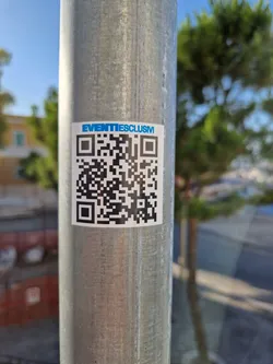A sticker featuring a QR code and the text 'EVENTIESCLUSM'. The sticker is affixed to a metal pole outdoors.