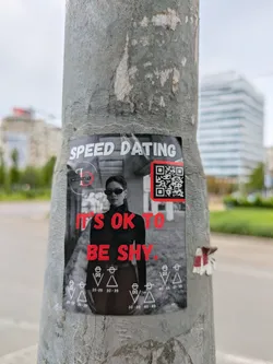 A black and white speed dating advertisement featuring a woman in sunglasses and the text 'IT'S OK TO BE SHY'. It contains a QR code.