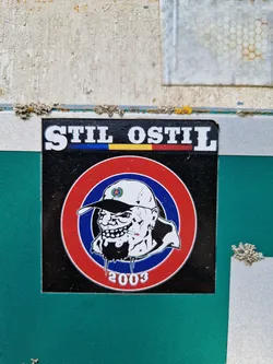 Street sticker A sticker featuring a caricature of a man with a goatee smoking a cigarette, within a red, white, and blue circular target. The text "Stil Ostil" is at the top, with "2003" at the bottom. The colors of the Romanian flag are present below the text. 