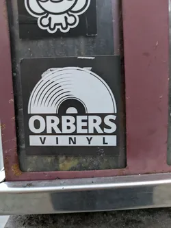 Street sticker A black and white sticker with a vinyl record illustration at the top and the text "ORBERS VINYL" at the bottom. The sticker is rectangular and placed on a weathered surface.