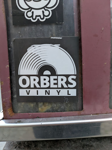 A black and white sticker with a vinyl record illustration at the top and the text "ORBERS VINYL" at the bottom. The sticker is rectangular and placed on a weathered surface.