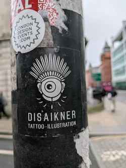 The central sticker is rectangular with a black background and white graphics. It features a stylized, wide-open eye with a prominent pupil and iris, topped by a radiating, spiky or feathered headdress design. Below the eye, several small, elongated teardrop or drip shapes are depicted. The overall aesthetic is bold and graphic. Below the illustration, the name 'DISAIKNER' is written in large, white, sans-serif capital letters, followed by 'TATTOO-ILLUSTRATION' in smaller, similar font. The sticker shows signs of wear, including scratches and some dirt.