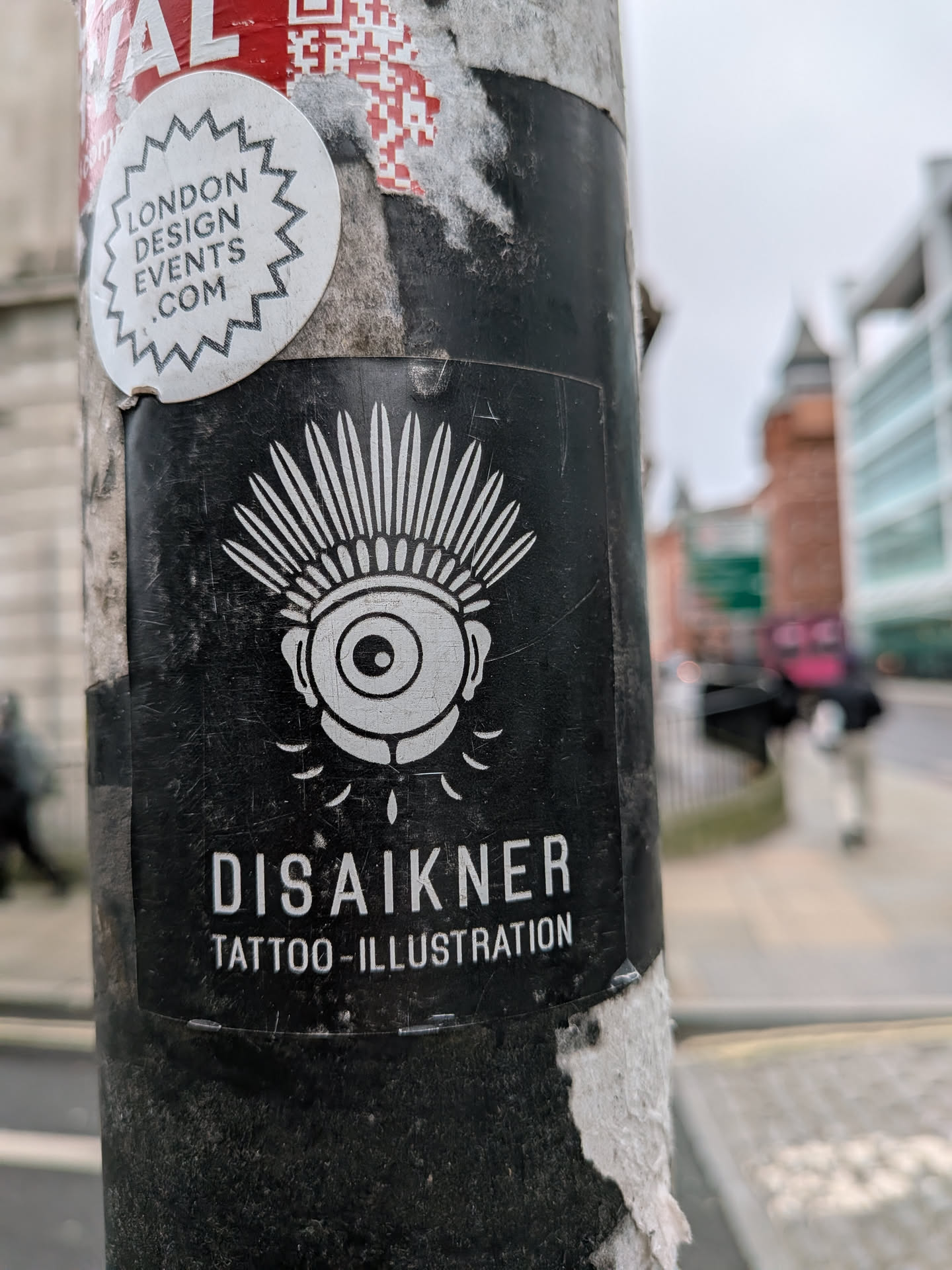 The central sticker is rectangular with a black background and white graphics. It features a stylized, wide-open eye with a prominent pupil and iris, topped by a radiating, spiky or feathered headdress design. Below the eye, several small, elongated teardrop or drip shapes are depicted. The overall aesthetic is bold and graphic. Below the illustration, the name 'DISAIKNER' is written in large, white, sans-serif capital letters, followed by 'TATTOO-ILLUSTRATION' in smaller, similar font. The sticker shows signs of wear, including scratches and some dirt.