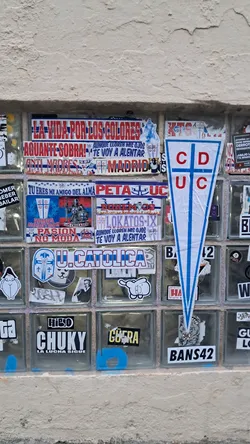 Street sticker The central sticker is a large, white pennant-shaped emblem with a prominent blue border and two vertical blue stripes forming a cross. Inside the white space, the letters 'CD' are stacked vertically on the left side of the cross, and 'UC' are stacked vertically on the right side, all in bold red capital letters. The sticker shows some minor creases, particularly towards the bottom, indicating it is a physical adhesive item.