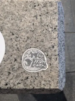 Street sticker A small, circular die-cut sticker on a granite surface. It features a white line-art illustration of a planet resembling Earth on the left, and the hand-written text 'FAR AWAY FROM EARTH' on the right, set against a dark background with a white border.