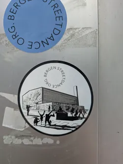 The central sticker is circular, with a black border and a white background. It depicts an urban setting in black and white, featuring a large, modern building on the left side, which has a detailed mural or graffiti covering part of its facade. In the foreground, several stylized figures are shown in dynamic street dance poses, including what appears to be breakdancing or b-boying. There is also a statue or monument-like structure and pigeons on the ground. The words "ORG. BERGEN STREETDANCE" are written in an arc around the top edge of the circle. The overall design suggests a focus on street culture and dance.