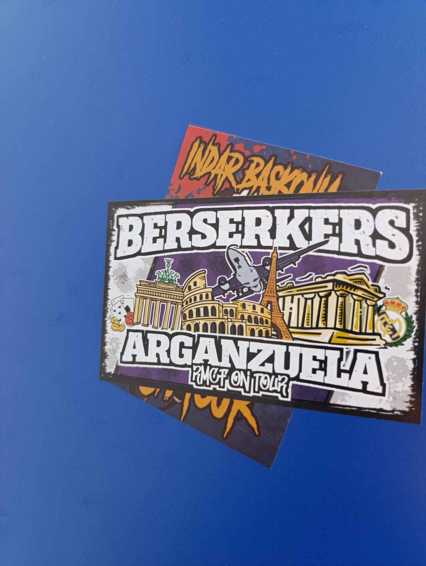 The sticker is rectangular with a distressed white and grey border, set against a vibrant purple background. At the top, 'BERSERKERS' is written in large, bold white letters with a black outline. Below, 'ARGANZUELA' is similarly styled. Underneath 'ARGANZUELA', smaller text reads 'RMGT ON TOUR'. The central design showcases a collage of iconic European landmarks in a cartoonish, golden-tan style: the Brandenburg Gate (with a green statue), the Colosseum, and the Eiffel Tower. A classical temple-like building is also present on the right. A grey airplane flies above these structures. To the left of the Brandenburg Gate, there are playing cards (Ace of Spades, Ace of Hearts), red dice, and gold coins. On the far right, the Real Madrid C.F. logo, featuring a crown and intertwined letters within a green laurel wreath, is prominently displayed.