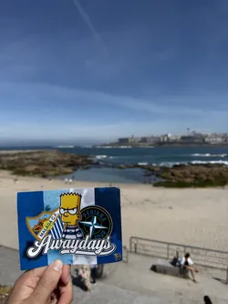 Street sticker A rectangular sticker featuring a background of vertical stripes in alternating shades of blue and white. Centered on the sticker is Bart Simpson, depicted with a stern expression, wearing a blue and white striped shirt. To Bart's left is the shield-shaped crest of Málaga C.F., which includes a castle, a lion, and a football, with 'MÁLAGA C.F.' written on a banner. To Bart's right is a circular emblem with a compass rose design, featuring the text 'PERIANA' at the top and 'TOUR' at the bottom. The word 'Awaydays' is prominently displayed in a large, stylized white script with a black outline across the lower half of the sticker. A tiny, less distinct cartoon character holding a flag is visible in the bottom right corner.