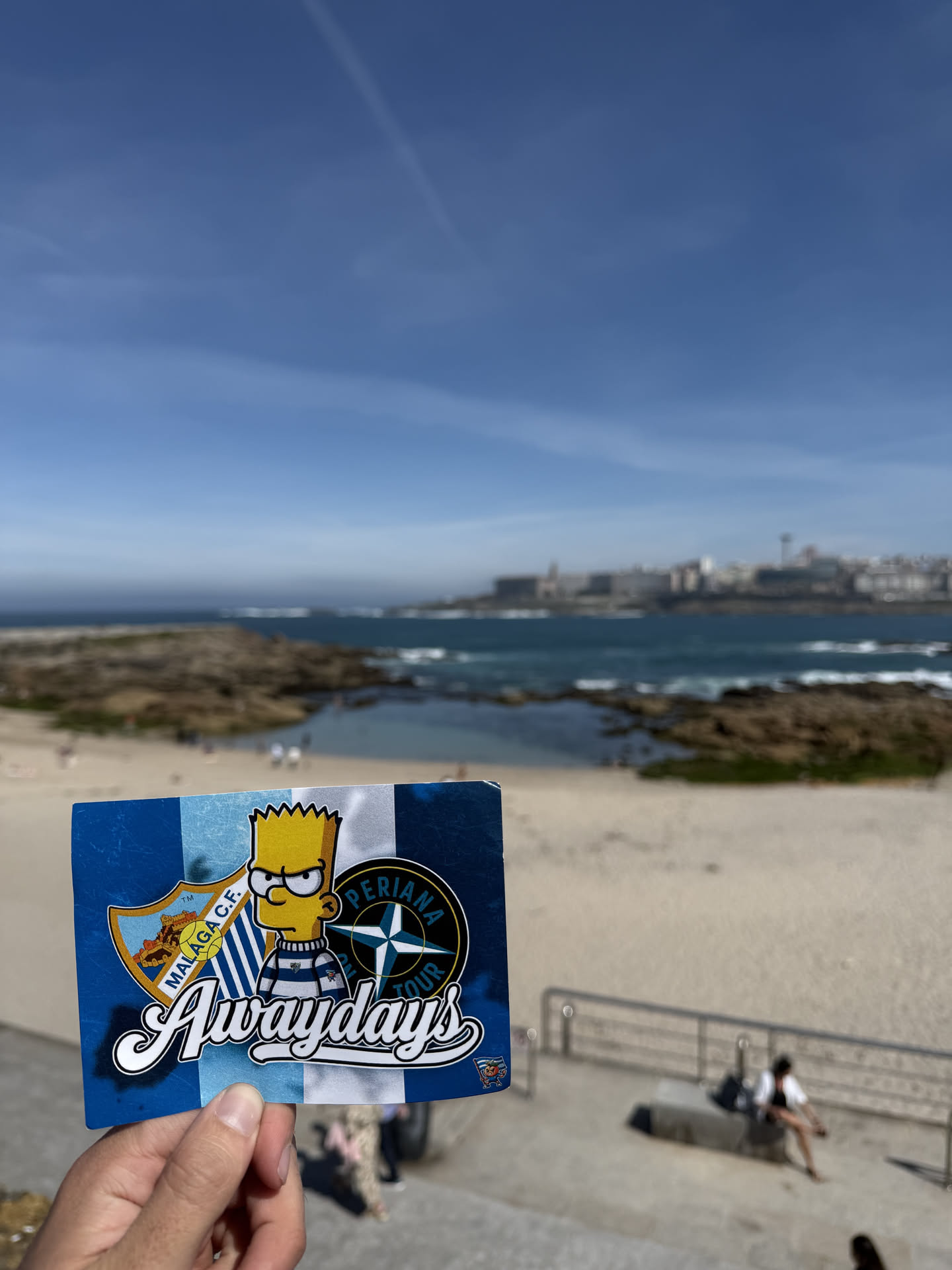 A rectangular sticker featuring a background of vertical stripes in alternating shades of blue and white. Centered on the sticker is Bart Simpson, depicted with a stern expression, wearing a blue and white striped shirt. To Bart's left is the shield-shaped crest of Málaga C.F., which includes a castle, a lion, and a football, with 'MÁLAGA C.F.' written on a banner. To Bart's right is a circular emblem with a compass rose design, featuring the text 'PERIANA' at the top and 'TOUR' at the bottom. The word 'Awaydays' is prominently displayed in a large, stylized white script with a black outline across the lower half of the sticker. A tiny, less distinct cartoon character holding a flag is visible in the bottom right corner.