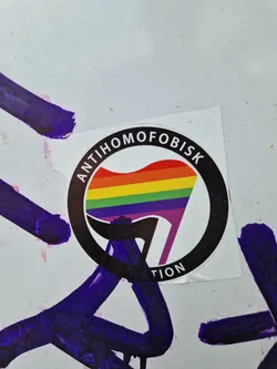 Street sticker A round sticker with a black border and a rainbow flag symbol inside. The text "Antihomofobisk Aktion" is written around the symbol.