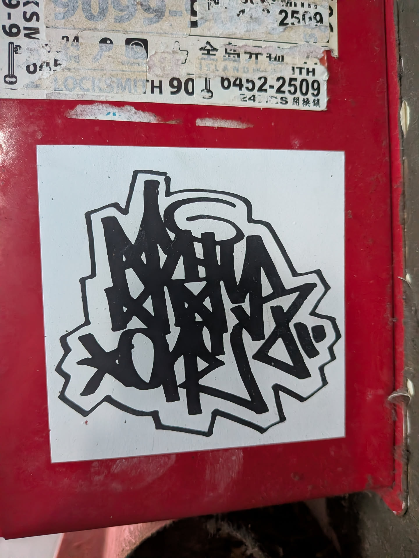 Square white sticker featuring a bold black graffiti handstyle tag with a thick outline, pasted on a red metal surface.
