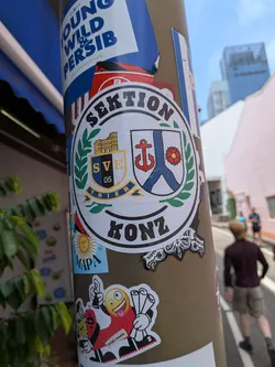 Street sticker A circular white sticker with black borders. It features the text 'SEKTION' at the top and 'KONZ' at the bottom. The center contains two shields framed by a green laurel wreath: the left shield represents SV Eintracht Trier 05 with the Porta Nigra monument, and the right shield displays the coat of arms of Konz, featuring a red anchor and a red rose.