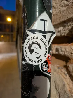 This sticker features a black and white portrait of a person wearing an eye patch. The portrait is encircled by text in a circular arrangement, reading "AMELAR LAPACA CABANYAL". The sticker is adhered to a pole, along with other stickers.