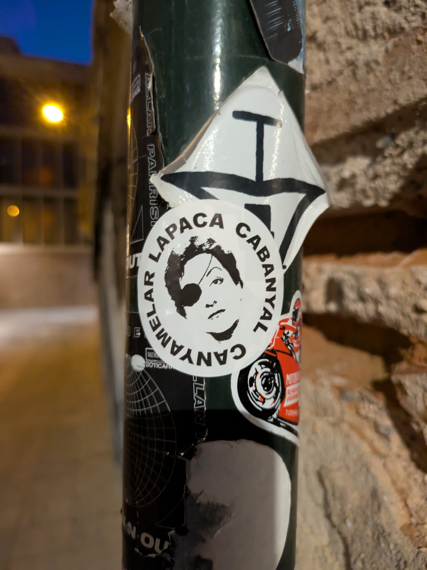 Street sticker This sticker features a black and white portrait of a person wearing an eye patch. The portrait is encircled by text in a circular arrangement, reading "AMELAR LAPACA CABANYAL". The sticker is adhered to a pole, along with other stickers.