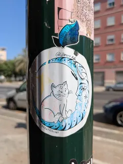 This circular sticker shows a white cat sitting comfortably on a moon with a human face. The moon is colored in shades of blue and white, while the cat has some light pink details. The sticker is placed on a dark green pole outdoors.