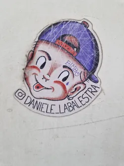 Street sticker This sticker features a charming cartoon drawing of a boy with a mischievous grin, sporting a purple baseball cap. His facial features are exaggerated and expressive. The word "BIMBO" is subtly written on his forehead, and a small tear is depicted beneath his eye. His tongue playfully protrudes from his mouth, adding to his playful character. The artist's Instagram handle, @Daniele_Labalestrra, is printed at the bottom of the sticker.