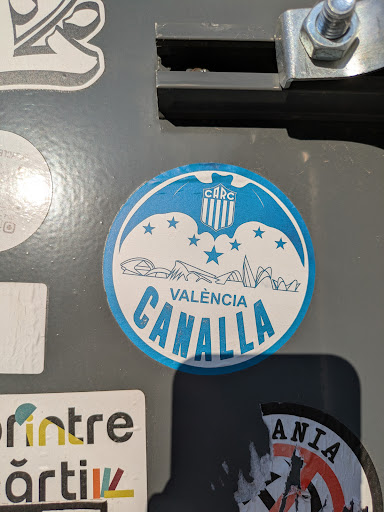 A round blue and white sticker features the text "VALENCIA CANALLA" in blue letters. Above the text is a stylized depiction of buildings and a logo with the letters "CARC". There are also several stars. The sticker appears to be adhered to a gray surface.