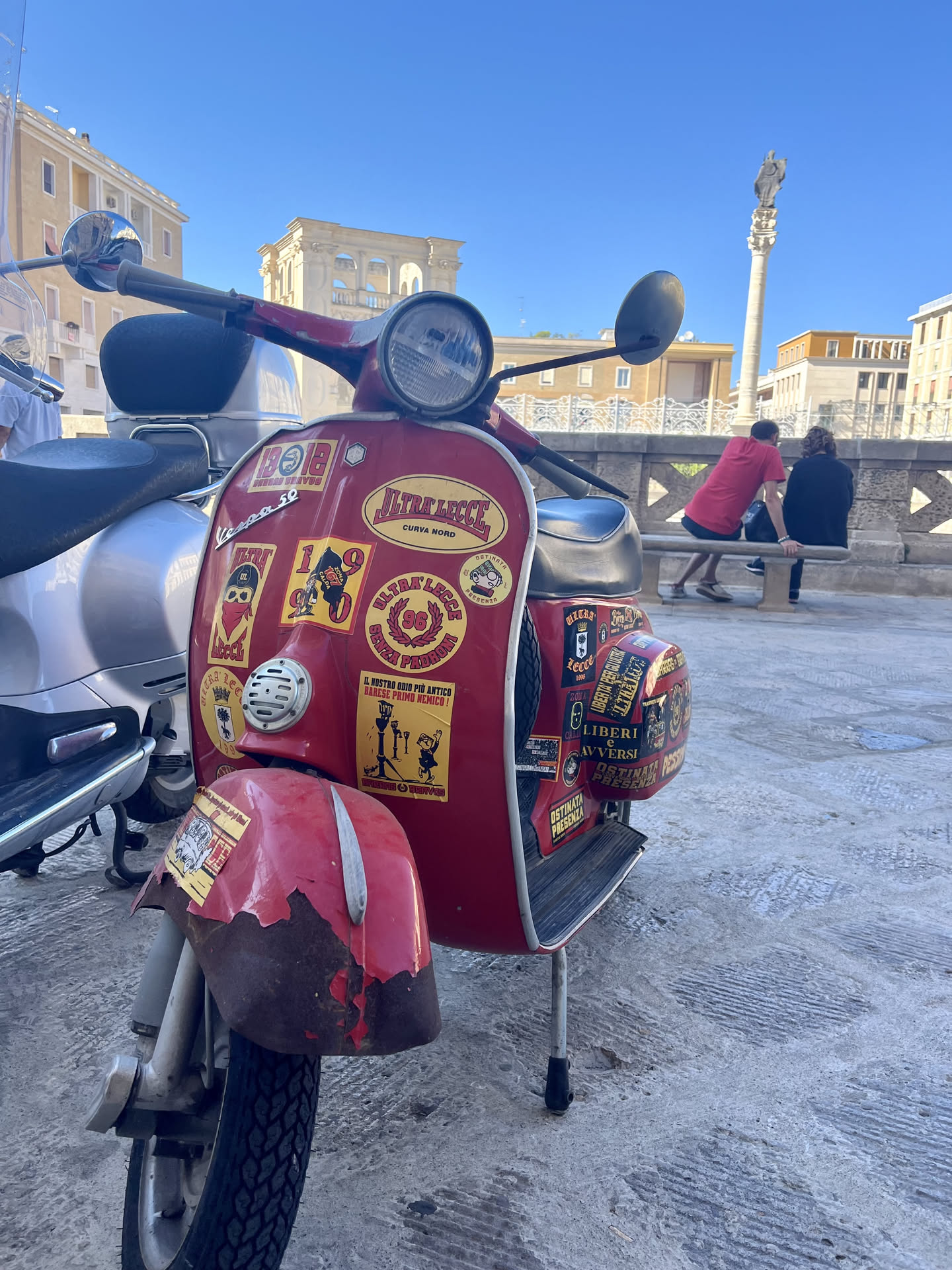 The central focus is a vintage red Vespa scooter, heavily adorned with numerous stickers, primarily on its front shield and right side panel. The stickers are mostly rectangular or oval, with a dominant color scheme of yellow backgrounds, black borders, and black or red text and graphics. Many stickers feature text related to "ULTRALECCE," "CURVA NORD," and "1908," indicating a strong affiliation with a football club's fan group. Common motifs include cartoon characters, some with bandanas or sunglasses, a character riding a scooter, and the Lecce coat of arms (a tree and a star). Slogans like "IL NOSTRO ODIO PIÙ ANTICO BARESE PRIMO NEMICO!" and "LIBERI E AVVERSI" convey a defiant and rivalrous tone. The stickers show varying degrees of wear, some peeling at the edges, adding to the scooter's character. The overall appearance is one of personalized expression and strong fan identity.