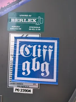 Street sticker The sticker is predominantly blue with a white border. Centered within the blue area are large, white letters in a gothic or old English font. The top line reads 'Cliff' and the bottom line reads 'gbg'. The sticker appears to be slightly creased and worn, suggesting it has been applied for some time. The overall aesthetic is striking due to the high contrast between the colors and the ornate font.