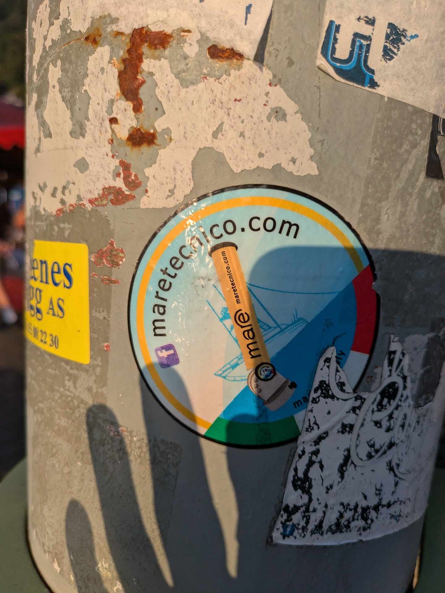 The sticker is round, prominently displaying 'maretecnico.com' along its upper edge. Its background is a light blue, likely representing the sea or sky, with faint outlines of what appears to be maritime vessels or structures. A brown, elongated object, resembling a pencil or a specialized tool, diagonally crosses the center, bearing the text 'mare @ maretecnico.com'. The bottom section of the sticker incorporates red and green colors, creating a subtle nod to the Italian flag, while a yellow arc frames the top. A Facebook social media icon is visible on the left side of the sticker. It is affixed to a heavily rusted and paint-peeling pole.
