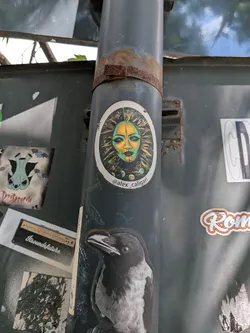 Street sticker An oval sticker depicting a stylized, mask-like face in green and yellow tones. The face is set against a dark, textured sunburst pattern. The handle '@alex_caligari' is printed at the bottom.