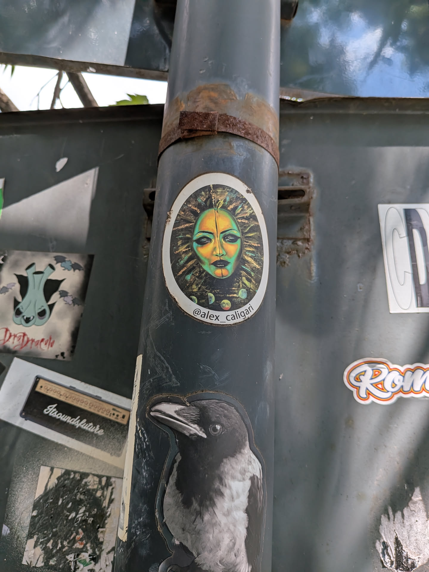 An oval sticker depicting a stylized, mask-like face in green and yellow tones. The face is set against a dark, textured sunburst pattern. The handle '@alex_caligari' is printed at the bottom.