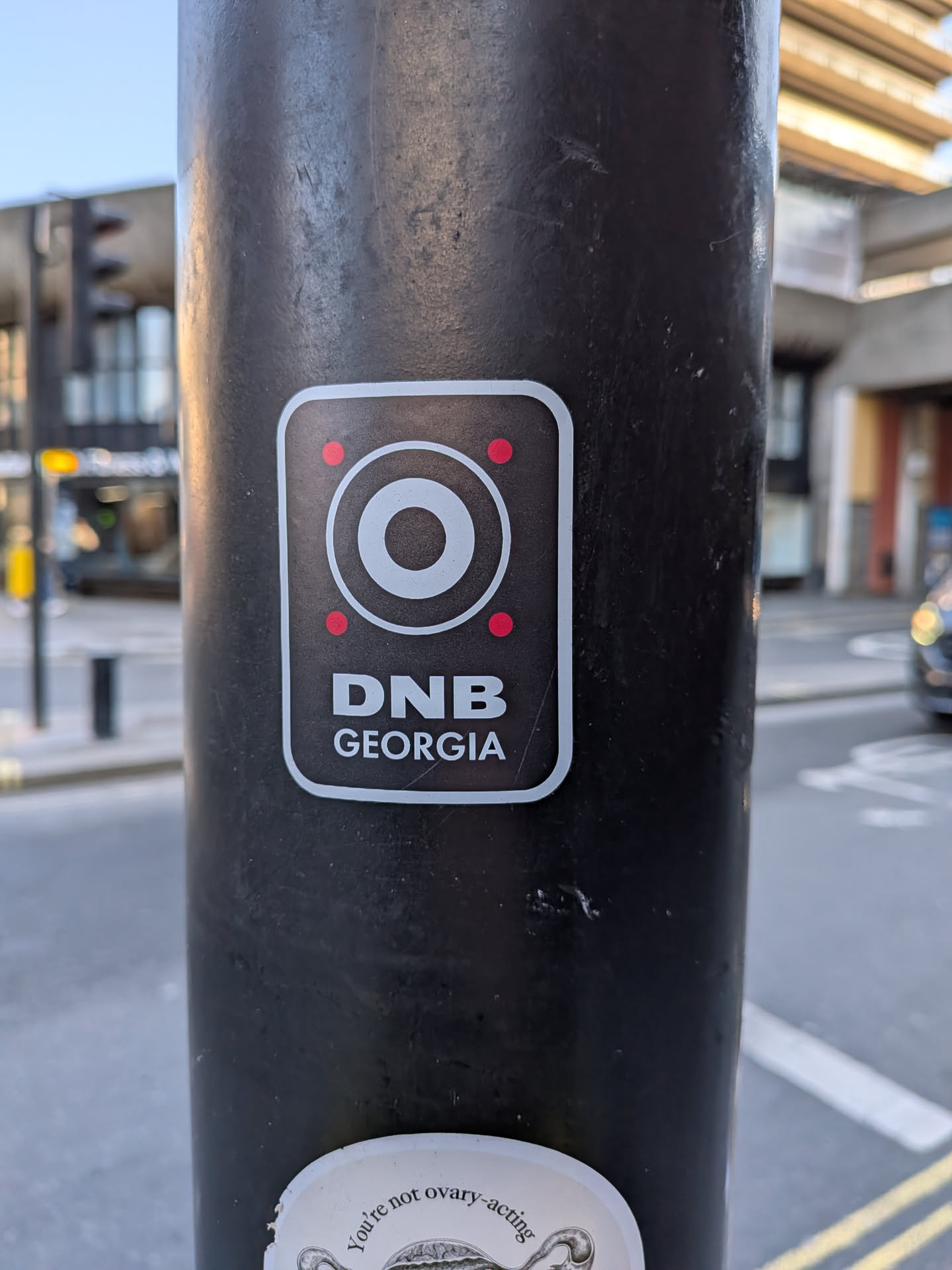 The sticker is rectangular with rounded corners, featuring a black background and a thin light grey border. At the top, there's a large white circular icon with a smaller concentric circle inside, resembling a speaker or audio output. Four small red dots are symmetrically placed around this central icon, two on the left and two on the right. Below the icon, in bold white capital letters, is the text "DNB". Further below, in smaller white capital letters, is "GEORGIA".