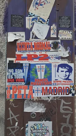 Street sticker A rectangular sticker featuring large, bold red block letters 'LP2' prominently displayed on a white background. Below the main 'LP2' text, in smaller blue lettering, it reads 'De la esquina a la cancha los parientes'. To the left of this smaller text, there is a small, partially obscured blue and white crest or logo, which appears to be a stylized 'CDUC' emblem. The sticker shows signs of wear, including minor discoloration and slight peeling at its edges, indicating its physical presence on a textured, weathered surface.