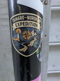 Street sticker The sticker is shield-shaped with a prominent gold border and background banners. At the top, a golden banner reads 'TAINARO - NORDKAPP', and below it, another banner says 'EXPEDITION'. The central design, set against a dark blue background, depicts a scenic adventure. On the left, a lighthouse stands by the sea under a large orange full moon or sun. In the center, a compass rose is clearly visible. To the right, there are mountains and evergreen trees, with a winding road leading towards a stylized blue globe at the bottom right. The overall design evokes themes of long-distance travel and exploration.