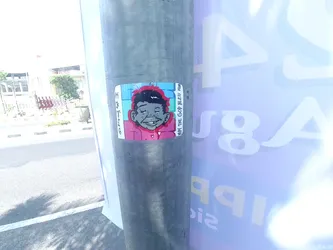 Street sticker A rectangular sticker on a metal pole featuring a grey-skinned cartoon boy smiling. He has a red outline, a pink shirt, and is set against a blue background. Text is written vertically on the left and right borders.