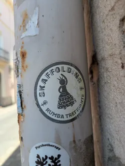 Street sticker A circular sticker on a pole featuring a stylized image of a dancing woman. The text around the edge reads "SKAFFOLDING RUMBA TREFIDANTE". The sticker is weathered.