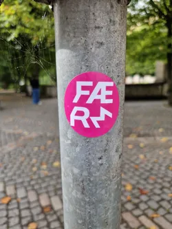 A round, pink sticker with the acronym FAER in white stylized lettering. The sticker is affixed to a weathered gray metal pole in an outdoor setting. The background is blurry, showing cobblestone pavement and some green trees.  The acronym FAER might be a brand name or abbreviation.