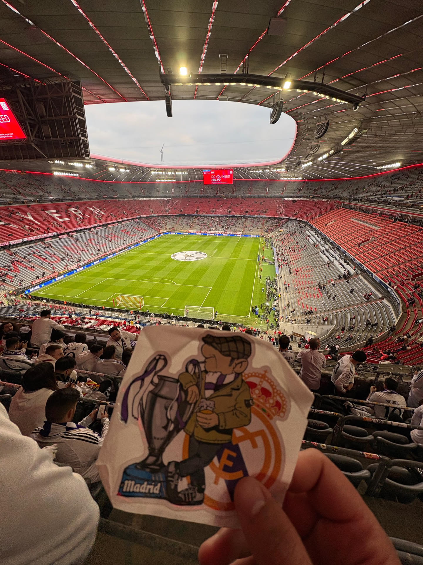A die-cut sticker featuring a cartoon illustration. On the left, a silver-grey UEFA Champions League trophy with purple ribbons sits on a blue base that reads 'Madrid'. To the right, a male cartoon character is depicted, wearing a brown jacket, a checkered flat cap, and a purple and white striped scarf. He holds a glass of what appears to be beer. Partially visible behind the character is the orange and gold crest of Real Madrid. The sticker has a white border and a slightly irregular shape.