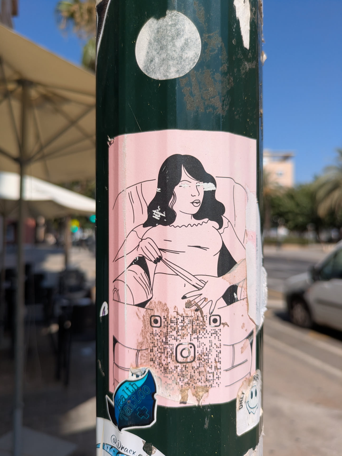 A pink sticker adhered to a green pole features a black and white line drawing of a woman sitting in a chair. The drawing style is simple and somewhat cartoonish.  A QR code is located towards the bottom of the sticker, along with what appears to be an Instagram logo. The sticker is peeling and shows signs of wear.